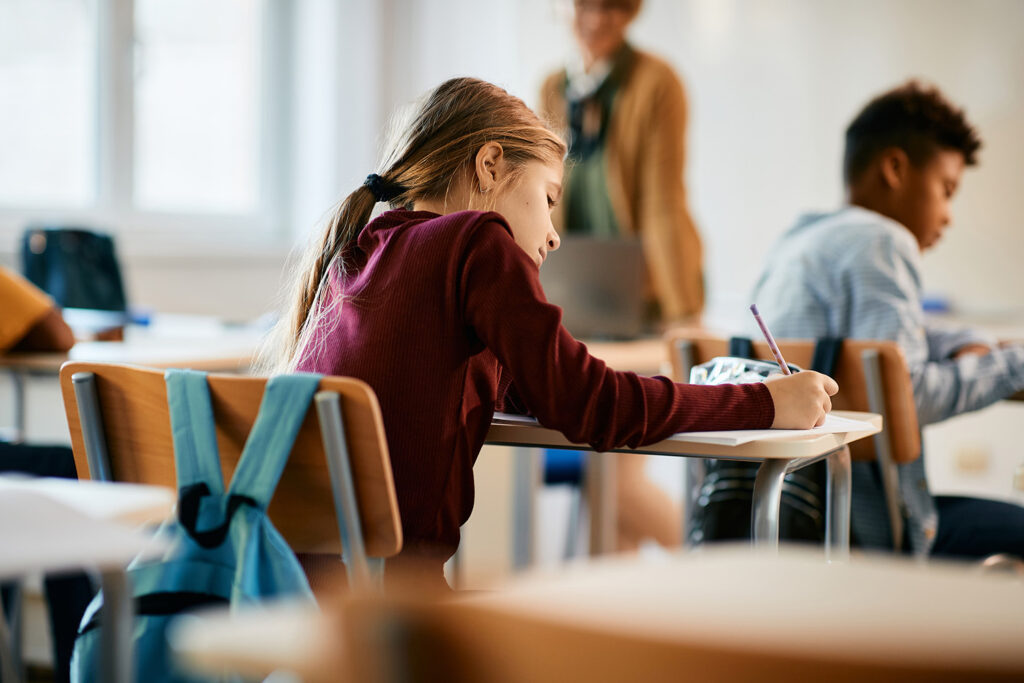 Ung skolflicka sitter vid sin skolbänk och skriver med en blyertspenna, fotad bakifrån med läraren i bakgrunden